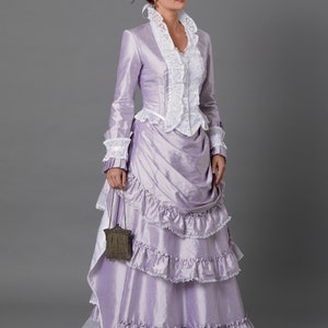 May include: A lavender Victorian-era dress with a matching hat. The dress features a fitted bodice with white lace trim, long sleeves, and a layered skirt with ruffles. A small, ornate purse hangs from the wrist. The hat is adorned with flowers and ribbons.