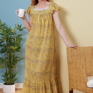 May include: A long, yellow nightdress with a small, dark pattern. The nightdress features short sleeves, a square neckline with lace trim, and a ruffled hem. A woman holds a white mug. The background is a mix of blue and yellow.