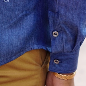 May include: Close-up of a blue denim shirt sleeve with two buttons. The cuff is buttoned. The shirt is paired with tan pants. The text "Simplicity Pattern 8427" is visible in the upper left corner.