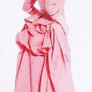 May include: A light pink Victorian-era dress with a fitted bodice, long sleeves, and a full skirt. The dress features a small, repeating pattern and is accessorized with a matching bonnet and scarf. The dress has a ruffled bustle.