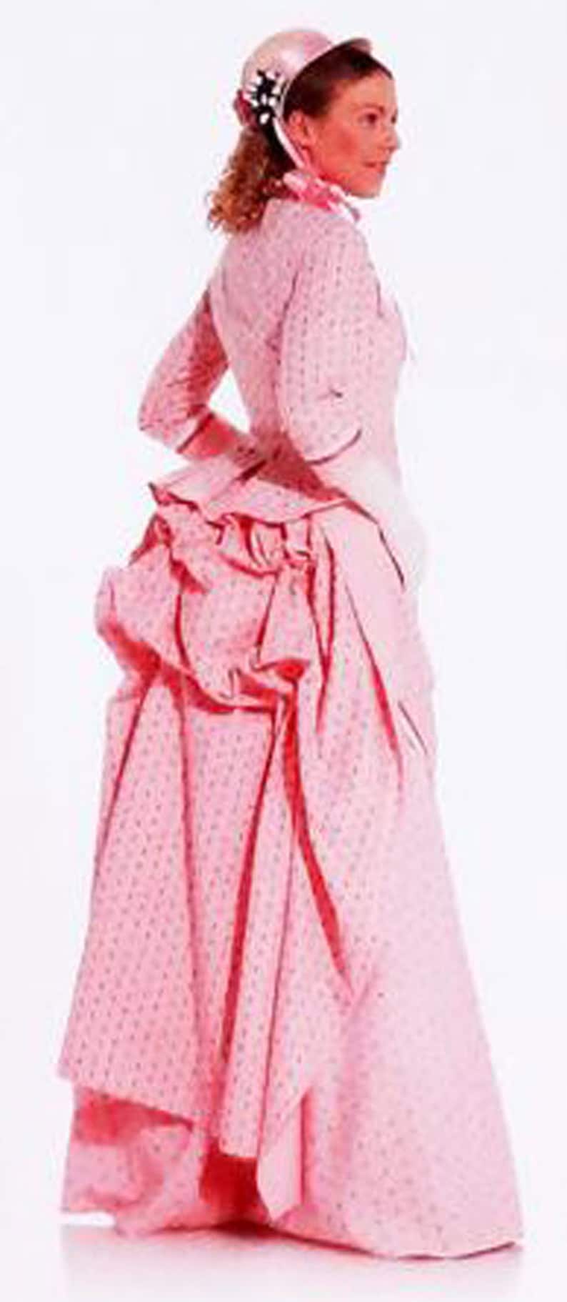 May include: A light pink Victorian-era dress with a fitted bodice, long sleeves, and a full skirt. The dress features a small, repeating pattern and is accessorized with a matching bonnet and scarf. The dress has a ruffled bustle.