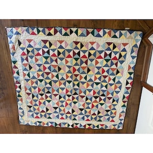 May include: A square quilt featuring a repeating pattern of small triangles in various colors including red, blue, yellow, green, and black. The quilt has a cream-colored border with a decorative stitched edge and a multi-colored triangle trim.