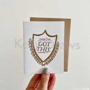 May include: A white card with a gold shield design that says "You've Got This." The shield is surrounded by a wreath of leaves.