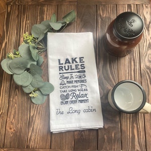 May include: A white kitchen towel with the text "LAKE RULES" in navy blue, along with illustrations and phrases like "Sleep In" and "Relax." A jar with a black lid and a white and black camping mug are also present.