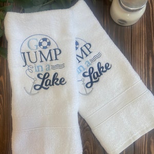 May include: Two white towels with blue and black embroidery. The towels say "Go Jump in a Lake" with a life preserver and anchor design.