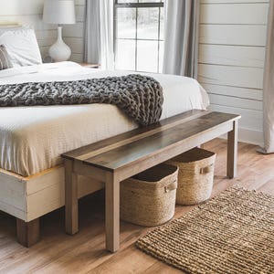 May include: A bedroom featuring a white wooden bed frame with a white duvet and a chunky dark gray knit throw blanket. The bed is adorned with several pillows, including one with black and white stripes. A wooden bench sits at the foot of the bed.