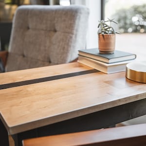 May include: A wooden side table with a black stripe across the top. The table has a black metal frame and is next to a gray armchair. There are three books and a small potted plant on the table.