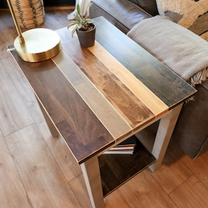 May include: A small wooden end table with a multi-toned wood top and a lower shelf. The table is next to a brown leather sofa with a throw blanket.