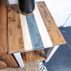 May include: A wooden side table with a multi-toned tabletop. The tabletop features alternating stripes of light and dark wood, with a blue stripe in the center. The table has a white base and a lower shelf. A dark vase sits on top.
