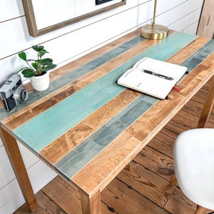 May include: A wooden desk with a multi-colored top featuring teal, gray, and brown planks. A small potted plant, a vintage camera, and an open notebook with a pen rest on the desk. The desk has four wooden legs.