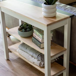 a white wooden side table with three shelves, displaying various items such as a potted plant, a stack of books, and a vase.