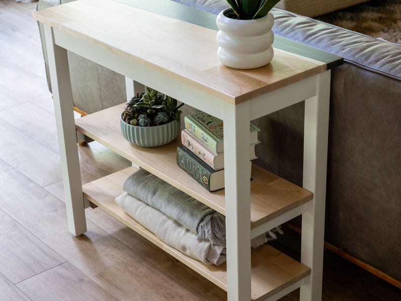 a white wooden side table with three shelves, displaying various items such as a potted plant, a stack of books, and a vase.
