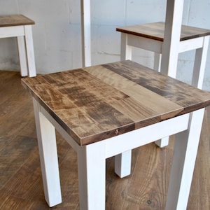 May include: A set of small wooden stools. The legs are painted white, and the tops are natural wood. The stools have a square shape. The room has a wooden floor and a light-colored wall. The stools are approximately 30 cm tall.