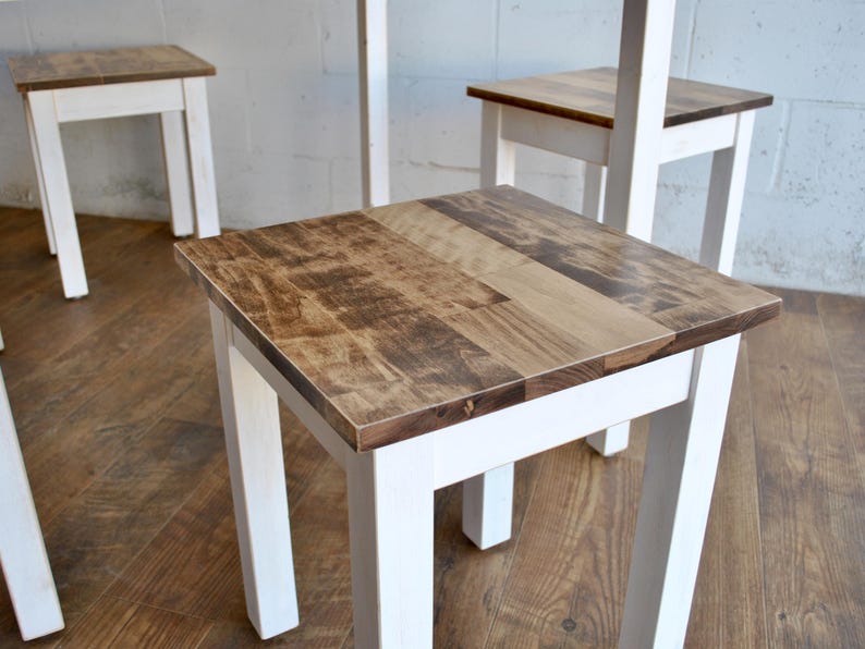 May include: A set of small wooden stools. The legs are painted white, and the tops are natural wood. The stools have a square shape. The room has a wooden floor and a light-colored wall. The stools are approximately 30 cm tall.