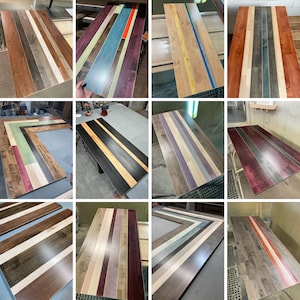 May include: A collection of rectangular wooden tabletops featuring various multi-colored striped patterns. Each tabletop is constructed from long, parallel wood planks finished in diverse stains and colors, including natural wood tones, deep reds, blues, purples, and blacks.