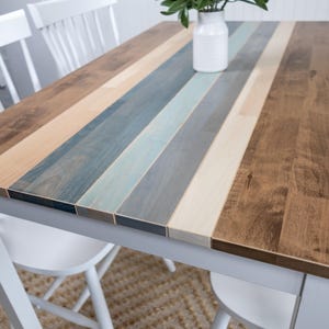 May include: A dining table with a white frame and a multi-toned wooden top. The tabletop has stripes of brown, beige, blue, and grey wood. A white vase with greenery is placed on the table, which is 120 cm long.