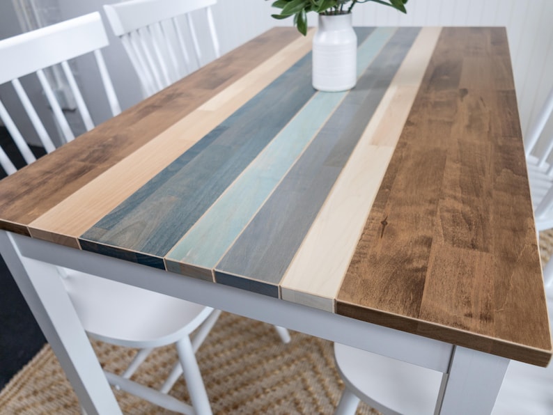 May include: A dining table with a white frame and a multi-toned wooden top. The tabletop has stripes of brown, beige, blue, and grey wood. A white vase with greenery is placed on the table, which is 120 cm long.