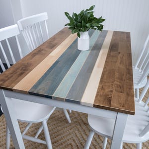 May include: A rectangular dining table with a multi-coloured wood plank top. The top features alternating stripes of brown, blue, and natural wood tones. Four white chairs surround the table. A white vase with greenery sits on the table.