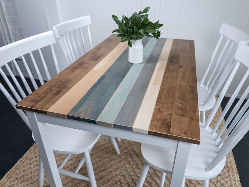 May include: A rectangular dining table with a multi-coloured wood plank top. The top features alternating stripes of brown, blue, and natural wood tones. Four white chairs surround the table. A white vase with greenery sits on the table.