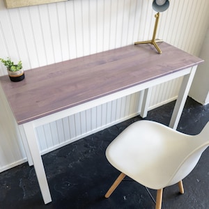 May include: A rectangular desk with a mauve-colored top and white legs. A small potted plant and a brass desk lamp sit on the desk. A white chair with wooden legs is in front of the desk.
