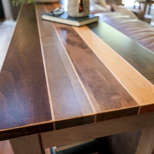 a wooden table with a multi-colored wood grain pattern, featuring a dark brown and light brown horizontal strip. The table is placed in a room with a leather couch and other furniture visible in the background.