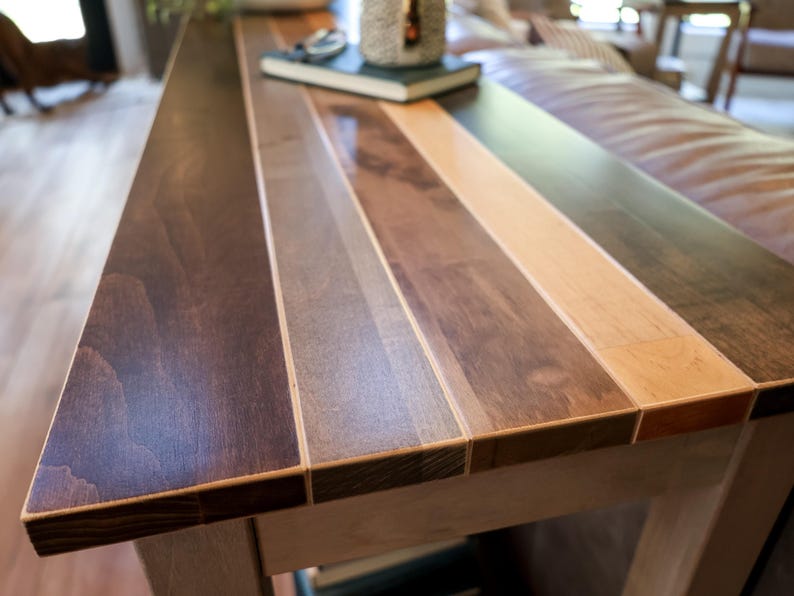 a wooden table with a multi-colored wood grain pattern, featuring a dark brown and light brown horizontal strip. The table is placed in a room with a leather couch and other furniture visible in the background.