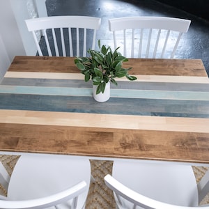 May include: A rectangular wooden table with a multi-coloured striped top, featuring shades of brown, blue, and grey. The table is supported by a white base and is paired with two white chairs. A small white vase with green leaves sits on the table.