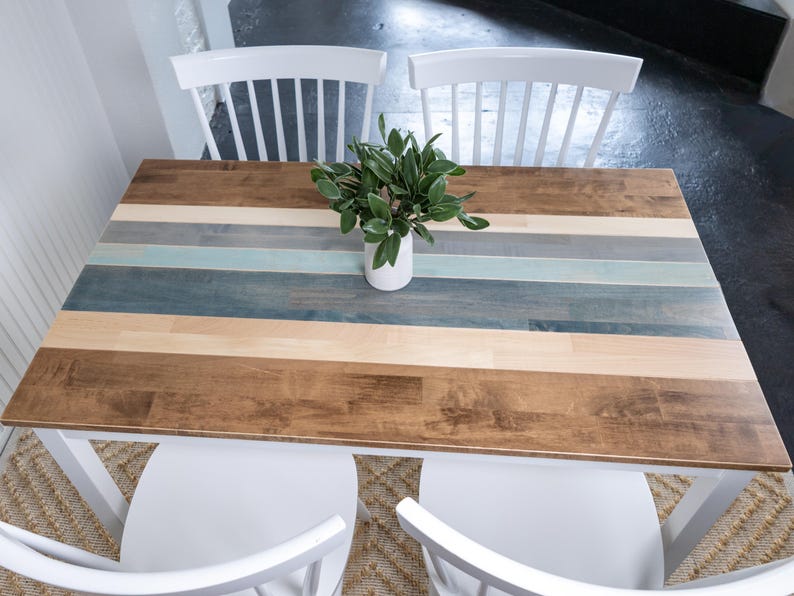 May include: A rectangular wooden table with a multi-coloured striped top, featuring shades of brown, blue, and grey. The table is supported by a white base and is paired with two white chairs. A small white vase with green leaves sits on the table.