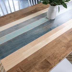 May include: A wooden table with a multi-coloured plank design. The table features alternating planks of light brown, medium brown, grey, and blue hues. A white vase with green leaves sits on the table. White chairs are visible.