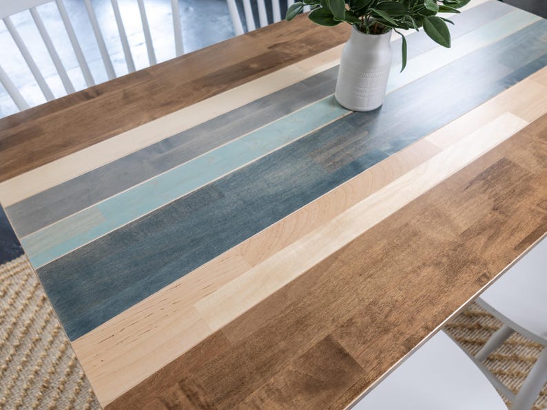 May include: A wooden table with a multi-coloured plank design. The table features alternating planks of light brown, medium brown, grey, and blue hues. A white vase with green leaves sits on the table. White chairs are visible.