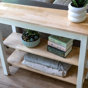 A wooden side table with three shelves, displaying various items such as a potted plant, a stack of books, and a decorative vase.