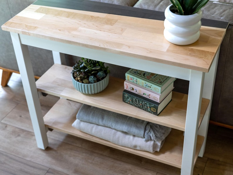 A wooden side table with three shelves, displaying various items such as a potted plant, a stack of books, and a decorative vase.
