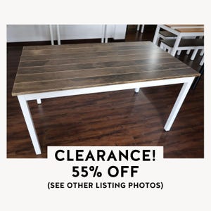 May include: A rectangular wooden table with a brown, slatted top and white legs. The table is on sale, with the text "CLEARANCE! 55% OFF" displayed below. The table appears to be approximately 60 inches long.
