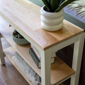 A wooden side table with a white vase of green plants and a stack of books on it, placed on a wooden floor in a living room setting.