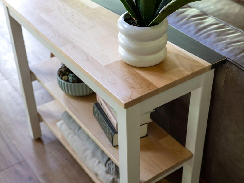 A wooden side table with a white vase of green plants and a stack of books on it, placed on a wooden floor in a living room setting.