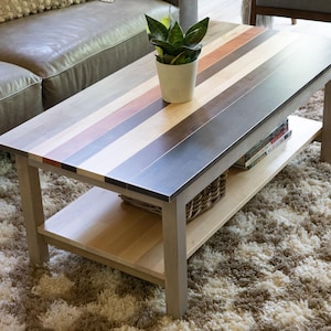 May include: A rectangular coffee table featuring a tabletop constructed from alternating horizontal planks of light, medium, and dark wood tones. The table includes four sturdy legs and a lower shelf for storage, with a small potted plant on top.