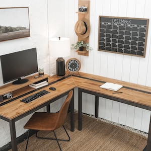 May include: A wooden L-shaped desk with a black stripe, a computer monitor, keyboard, and a brown leather chair. The desk is in a home office setting with a calendar, artwork, and a lamp.