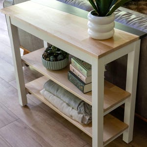 a white wooden side table with a shelf on the top, displaying various items such as a potted plant, a stack of books, and a bowl of decorative objects.
