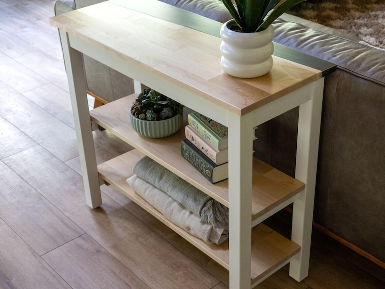 a white wooden side table with a shelf on the top, displaying various items such as a potted plant, a stack of books, and a bowl of decorative objects.