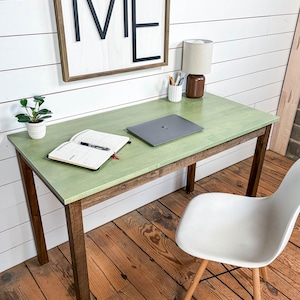 May include: A green desk with brown legs, a white chair, and a wooden sign that says "ME". A laptop, a notebook, a pen, a small potted plant, and a lamp are on the desk. The desk is in a room with white shiplap walls and wood flooring.