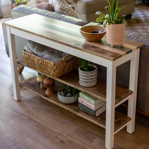 May include: A rectangular console table with a white frame and a natural wood top. It features two lower shelves holding a woven basket, a small potted plant, wooden mushroom figurines, a small bowl of succulents, and a stack of three books.