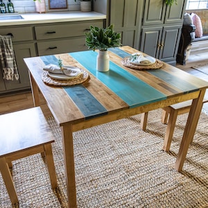 May include: A wooden dining table with a turquoise stripe down the center, flanked by natural wood tones. The table is set with plates, placemats, and a vase of greenery. A matching stool sits nearby, all on a woven rug.