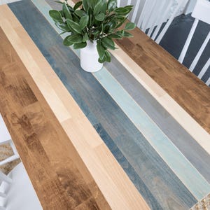 May include: A wooden table with a multi-coloured striped design. The table features alternating stripes of brown, beige, blue, and grey. A white vase with green leaves sits on the table. Two white chairs are visible.