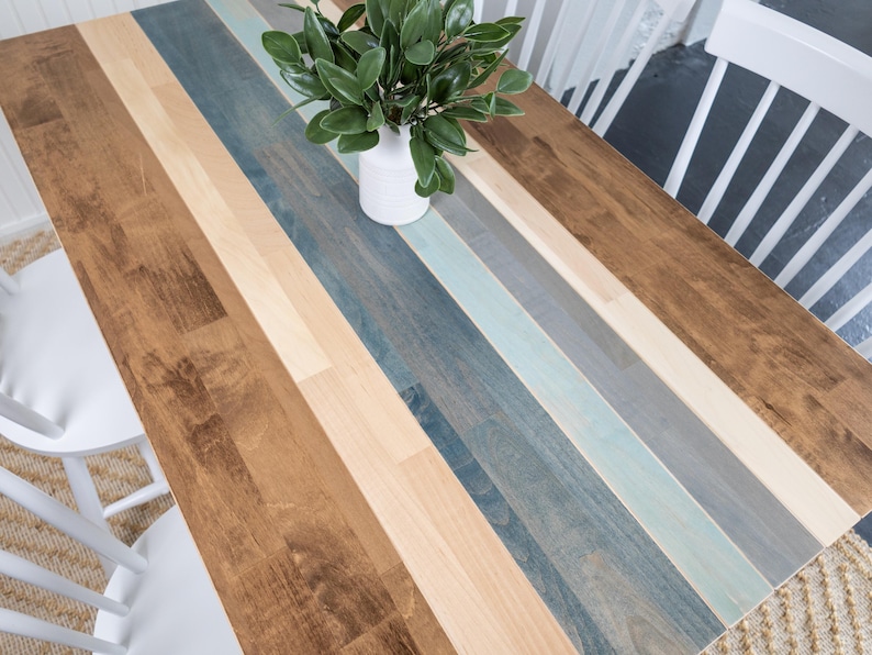 May include: A wooden table with a multi-coloured striped design. The table features alternating stripes of brown, beige, blue, and grey. A white vase with green leaves sits on the table. Two white chairs are visible.