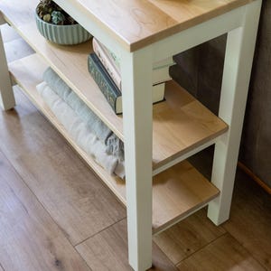 A white wooden side table with three shelves, holding various items such as books, a bowl, and a plant.