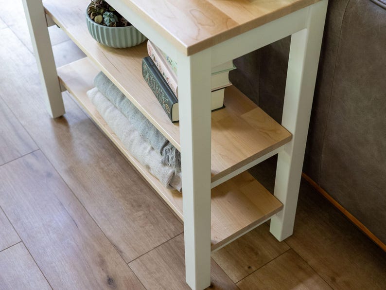 A white wooden side table with three shelves, holding various items such as books, a bowl, and a plant.