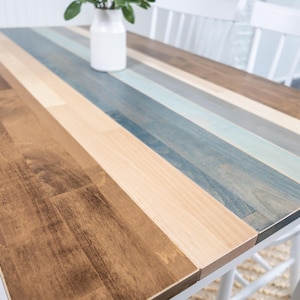 May include: A rectangular wooden table with a multi-coloured striped top. The stripes alternate between shades of brown, beige, and blue. The table is supported by white legs and is suitable for dining or as a decorative piece. The table measures approximately 152 cm long.