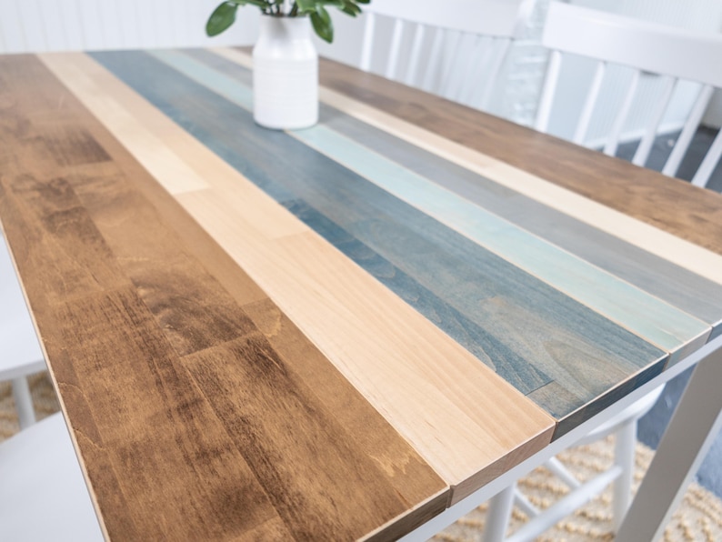 May include: A rectangular wooden table with a multi-coloured striped top. The stripes alternate between shades of brown, beige, and blue. The table is supported by white legs and is suitable for dining or as a decorative piece. The table measures approximately 152 cm long.