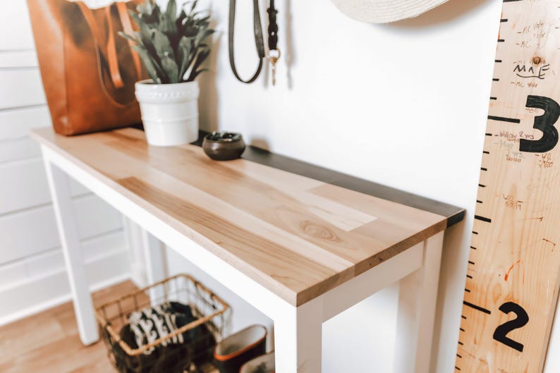 a wooden console table with a white top, a potted plant, a leather bag, a small bowl, and a metal ruler on the wall next to it.