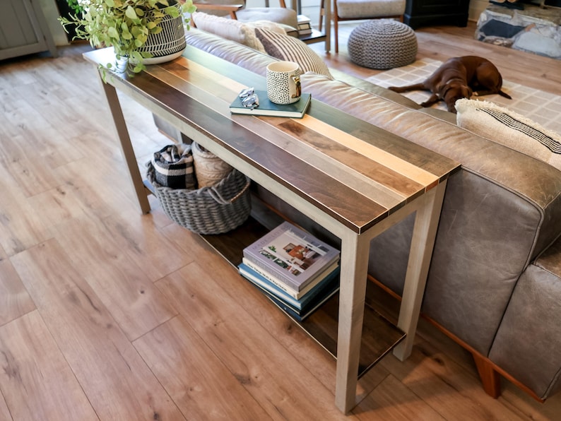 a cozy living room with a wooden console table, a gray sofa, and a brown dog resting on the floor. The table has a striped top and is decorated with various items, including a plant, a coffee cup, and a stack of books.